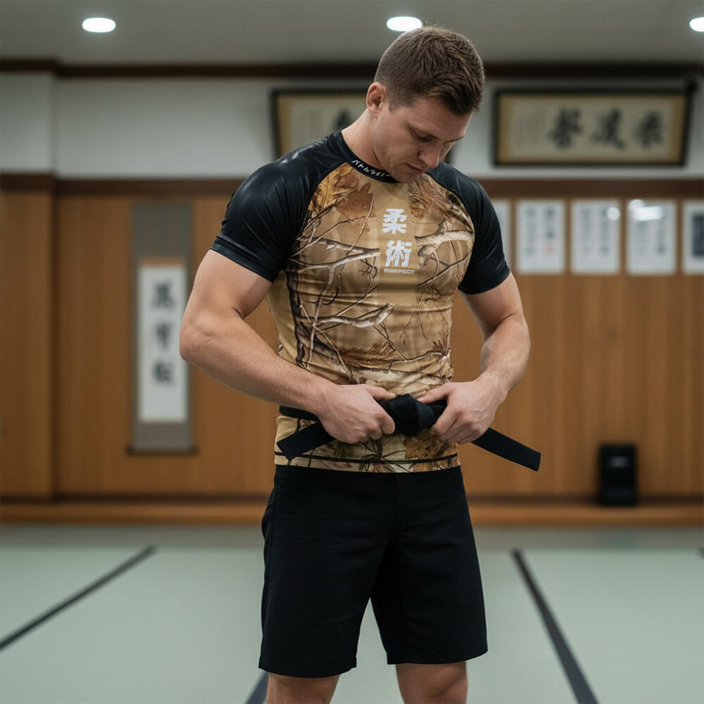 Male model wearing RISEPECT autumn yellow-brown short sleeve rash guard with Japanese jiu-jitsu lettering and tree branch and leaf pattern, tying his black jiu-jitsu belt in a gi training room.